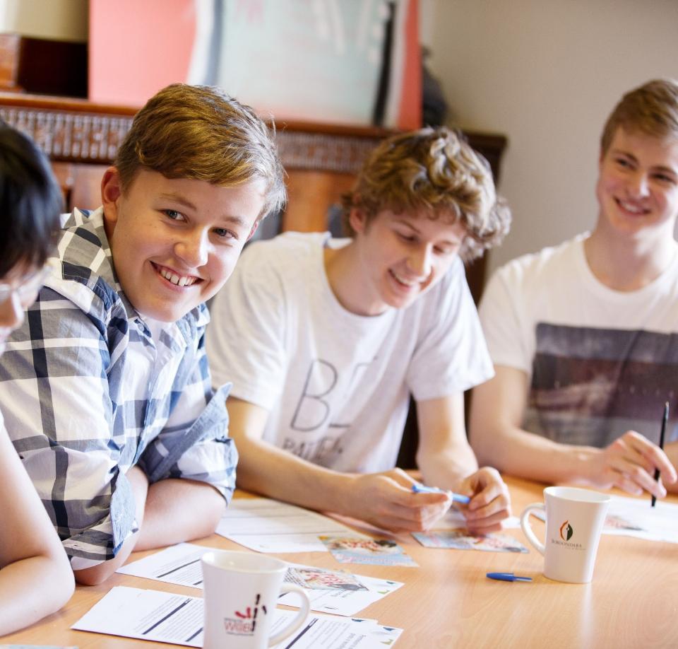 Four teenage boys sit at a table smiling at each other. They have paper forms in front of them and some are holding pens.