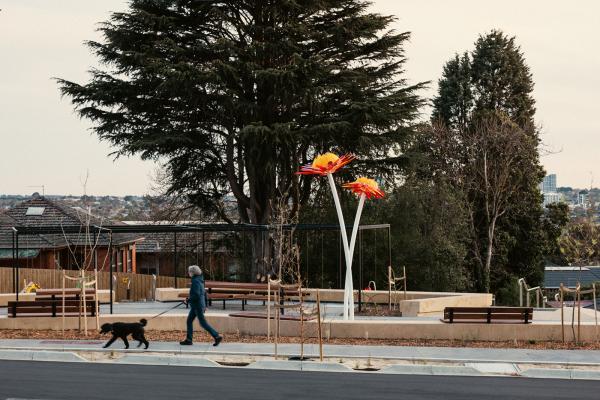 A woman walks her dog in front of a contemporary urban park with large trees in the background. At the centre of the park is a large sculpture which takes the form of two orange flowers on long stems.