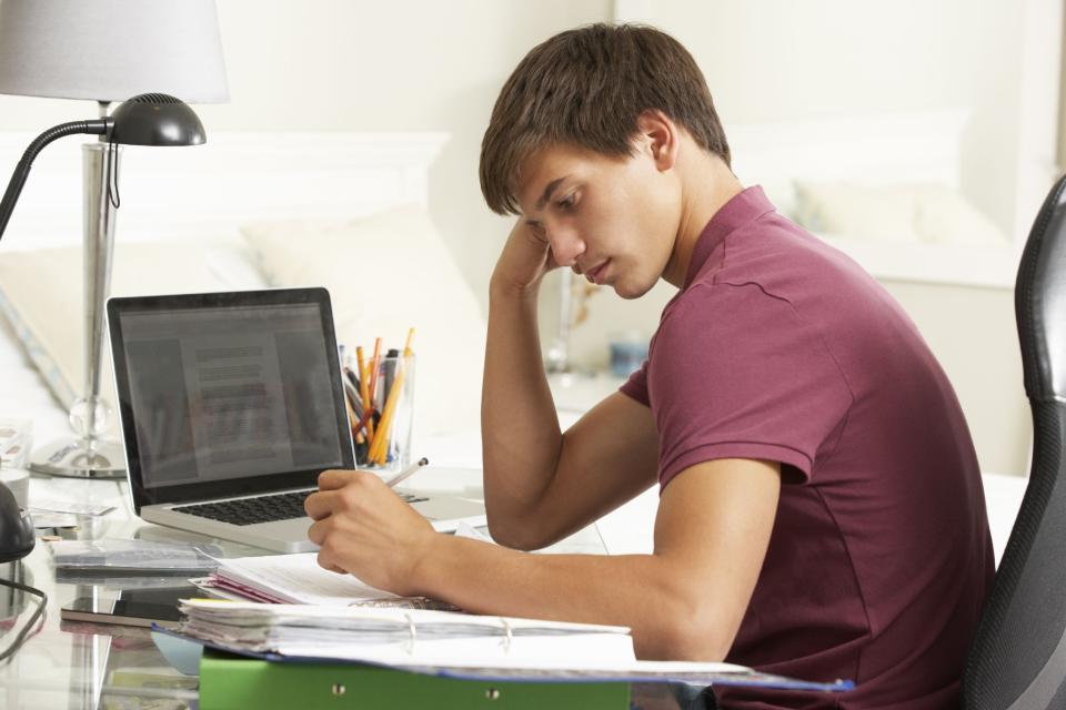 A teenage boy sits at a desk writing. A laptop is open beside him and he has his hand resting on the side of his face.