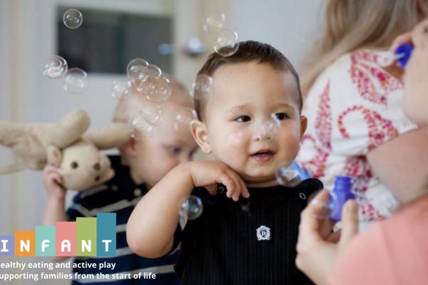 A smiling baby looks toward an adult who is blowing bubbles.