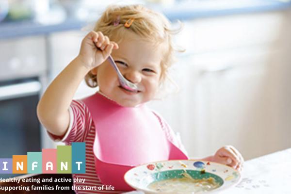 Smiling toddler wearing a pink bib eats with a spoon at a table, with a bowl of food in front. Text reads: “INFANT – Healthy eating and active play. Supporting families from the start of life.”