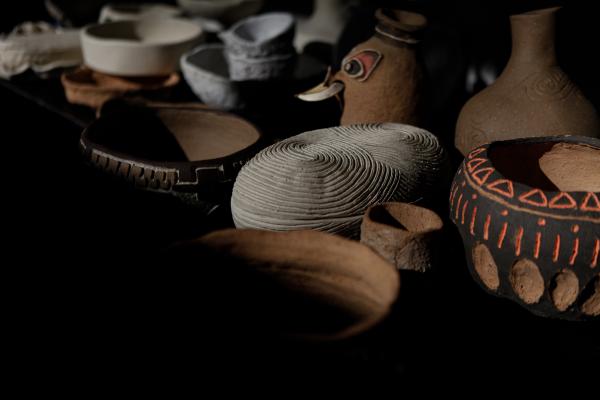 A collection of handcrafted pottery, including painted and textured clay vessels, bowls, and jars, displayed in soft lighting with shadows adding depth.