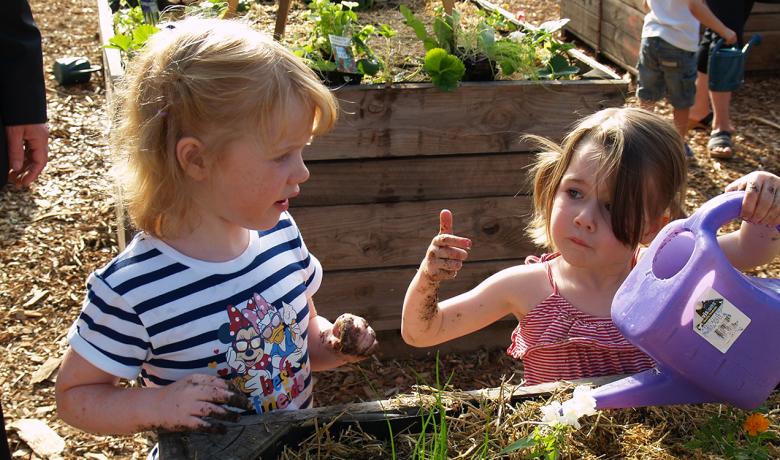 Two children gardening together in a raised garden bed. One holds a purple watering can while the other has soil on their hands. Green plants and garden beds are visible in the background.