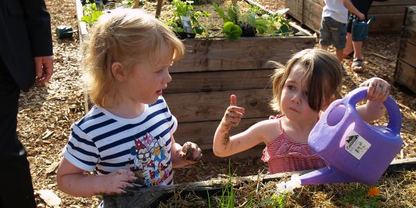 Two children gardening together in a raised garden bed. One holds a purple watering can while the other has soil on their hands. Green plants and garden beds are visible in the background.