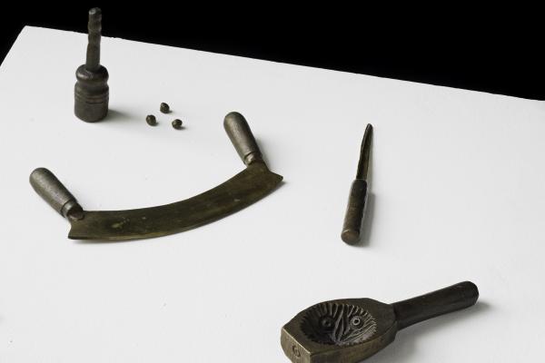 A collection of old metal tools displayed on a white surface, including a curved blade with handles, a metal mold with a handle, a pointed tool, a cylindrical piece and three bullets.