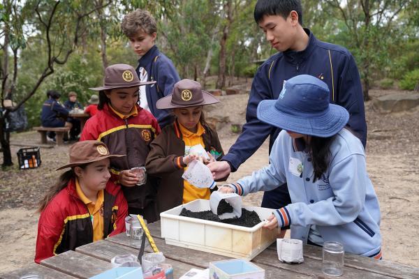 A group of kids of mixed ages and genders stand at a wooden table in a bushy environment. They are scooping dirt into plastic containers.