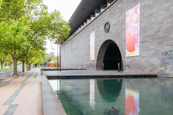 A wide paved footpath with large green Elm trees runs beside a modern grey brick building surrounded by a moat. 