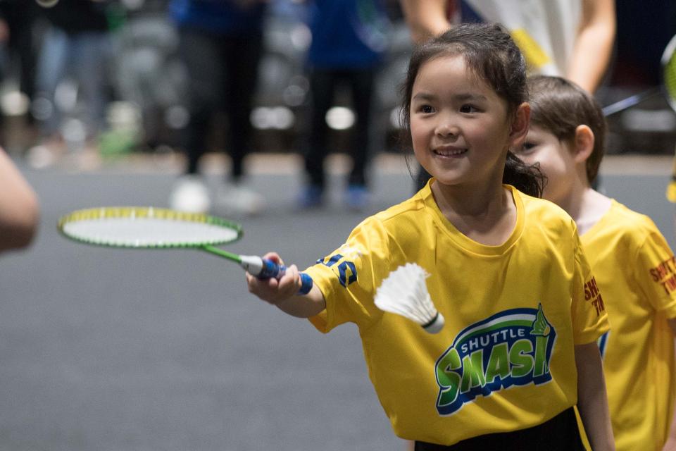 A young girl smiling and holding out a badminton racquet. She is wearing a yellow t-shirt with the words 'shuttle smash' on it. Another child is waiting behind her.