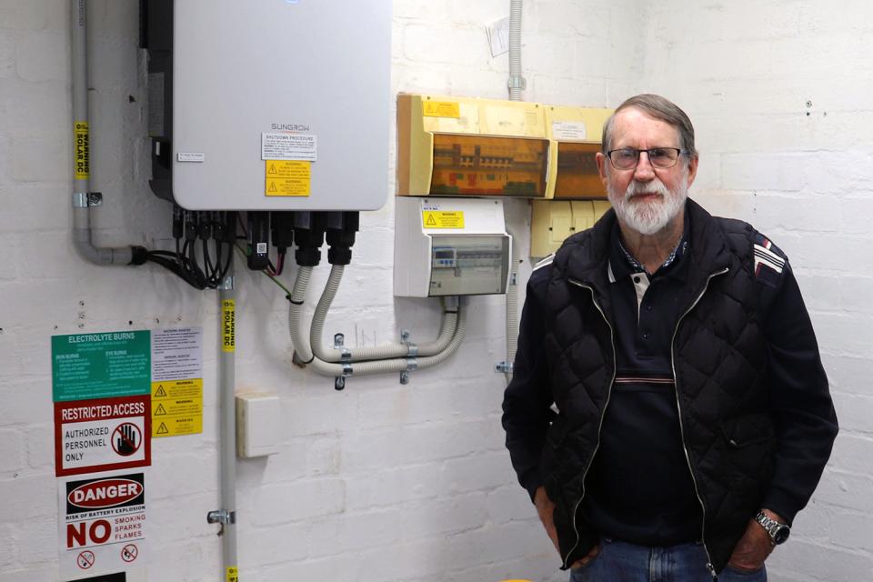 A man wearing glasses and a black jacket stands with his hands in his pockets. He is in a garage and a solar battery and a switchboard can be seen on the wall to his right.