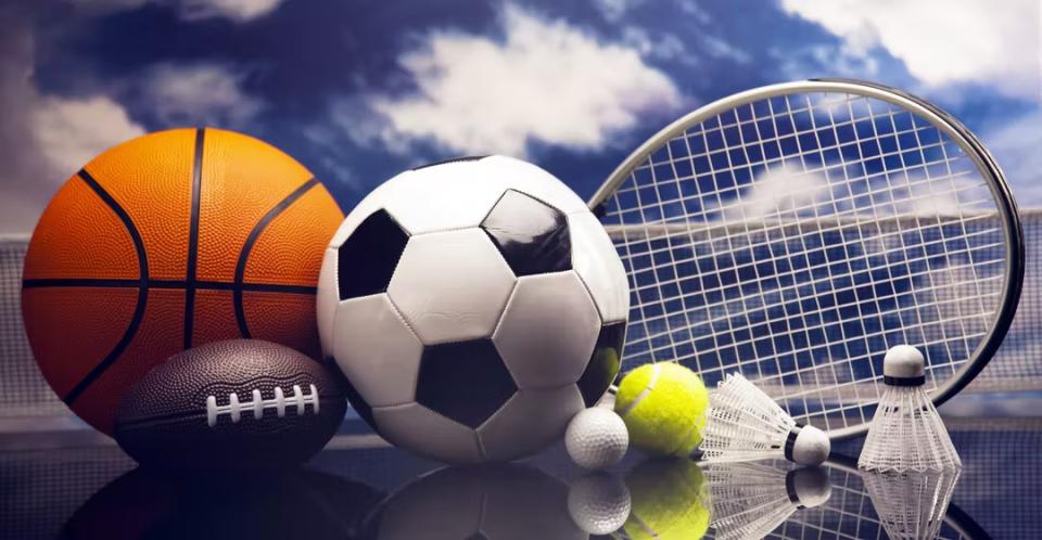 Basketball, soccer ball, NFL ball, tennis racquet and balls, and shuttlecock in foreground with blue sky and clouds in background