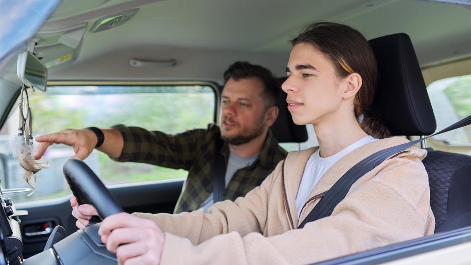 A middle-aged man sits beside a teenage boy who is driving a car. The older man is pointing towards the windscreen.