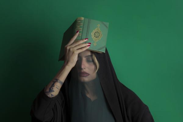 A woman dressed in black with a sheer veil sits against a green background. She holds a book with a gold emblem on her head and a green beaded rosary in one hand, with a white flower nearby.