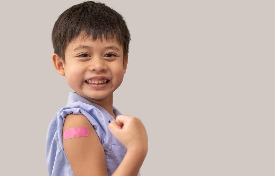 Smiling young boy flexes arm to show pink bandage after vaccination, he's wearing light blue shirt.