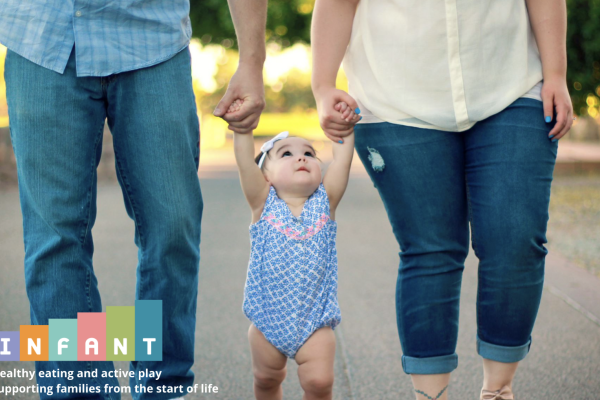 baby in between parents, holding their hands as they walk. Only the parents' legs are in view.